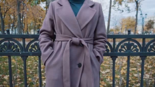 Ein Mädchen in fliederfarbenem Mantel mit Gürtel steht am Zaun des Parks. Mantel aus nächster Nähe — Stockvideo