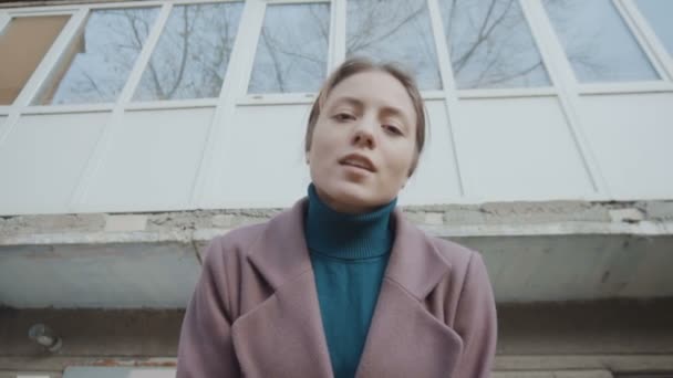 Девушка в фиолетовом пальто стоит на фоне старого здания и смотрит. Рамка удаляется от девушки — стоковое видео