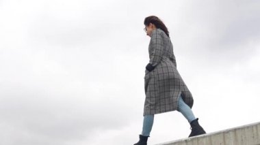 Genç bir kadın beton bir çitin üzerinde yürüyor. Palto giyen kız.