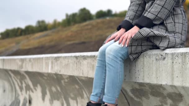 Young woman sits on a concrete fence in the wind and shakes her legs — Stock Video