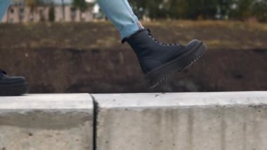 Bir kadın büyük siyah botlarla beton bir çit boyunca yürür. Kapat.