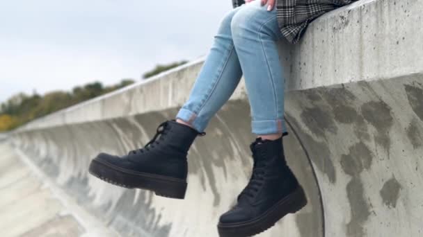 Young woman sits on a concrete fence in the wind and shakes her legs — Stock Video