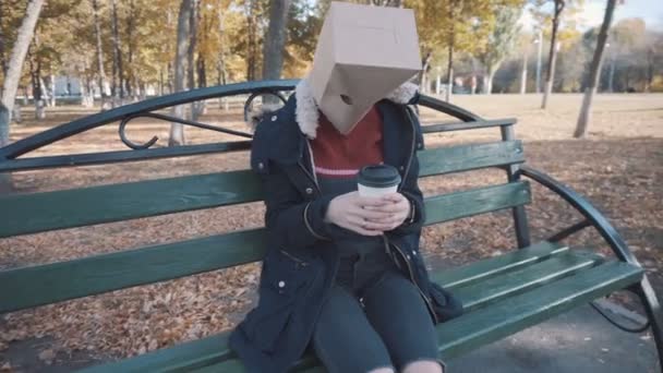 La chica con el paquete en la cabeza sentada en un banco con una taza de café. Chica en una chaqueta — Vídeos de Stock