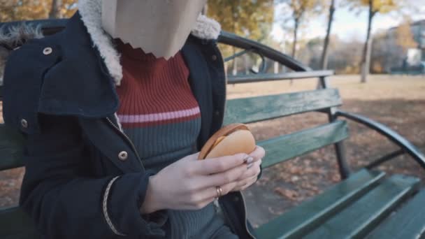 La chica con el paquete en la cabeza está sentada en un banco con una hamburguesa con queso. Chica en una chaqueta — Vídeos de Stock