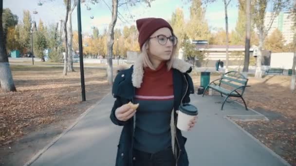 Flicka går i parken och äter en hamburgare och dricker kaffe.Flicka i jacka och hatt — Stockvideo