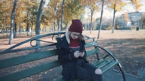 Dívka sedí na lavičce v parku s telefonem. Dívka v bundě a klobouku — Stock video