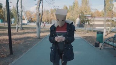 Kafasında bir paket olan bir kız elinde bir fincan kahveyle ağır çekimde yürür. Ceketli kız.