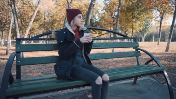 A lány kávét iszik egy padon a parkban. Lány zakóban és kalapban — Stock videók