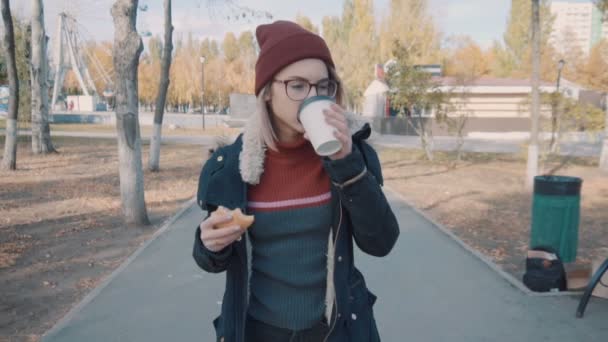 Chica camina en el parque y beber café lentamente. Chica en una chaqueta y sombrero — Vídeo de stock