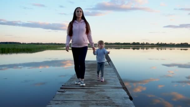 Anya és a kislánya sétálnak a hídon naplementekor. Van egy tó a híd körül. Felhők tükröződnek a tóban — Stock videók