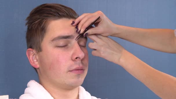 A young man plucked eyebrows in a spa salon. Woman tweezers pulls out eyebrows. A man in a white coat.. Head close up — Stock Video