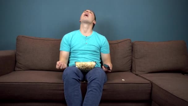 Un joven está viendo televisión y comiendo palomitas de maíz. El tipo atrapa palomitas con la boca. Sentado en el sofá. La vista desde el televisor. Mirando a la cámara — Vídeo de stock