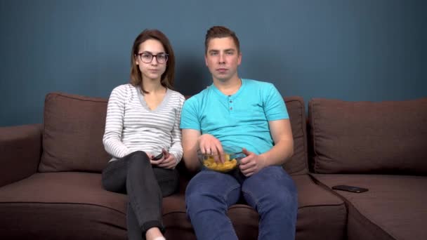 Ένας άντρας και μια γυναίκα βλέπουν τηλεόραση. Ένα νεαρό ζευγάρι κάθεται στον καναπέ και τρώει πατατάκια. Πιάτο γυάλινο με πατατάκια στα χέρια — Αρχείο Βίντεο