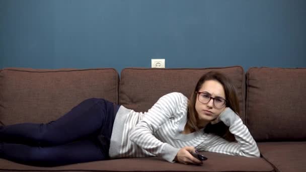 Μια νεαρή γυναίκα με γυαλιά βλέπει τηλεόραση. Κορίτσι ξαπλωμένο στον καναπέ — Αρχείο Βίντεο