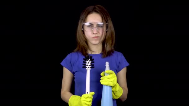 Eine junge Frau kratzt sich mit einer Klobürste am Kopf. Frau in Schutzbrille und Handschuhen mit Werkzeug für die Toilettenreinigung. Mädchen hält Klobürste und Spray. auf schwarzem Hintergrund — Stockvideo