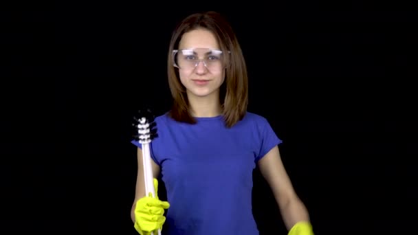 Una joven está llegando a un baño con un cepillo de inodoro y mostrando bíceps. Mujer con gafas de seguridad y guantes con herramientas para limpiar el inodoro. La chica sostiene un cepillo de baño. Sobre un fondo negro — Vídeos de Stock