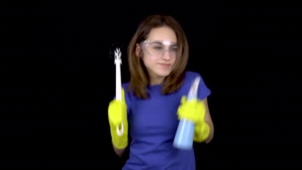 En ung kvinna dansar med toalettborste och spray. Kvinna i skyddsglasögon och handskar med verktyg för att rengöra toaletten. Flickan håller en toalettborste och vattenspray. På en svart bakgrund — Stockvideo