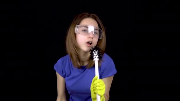 A young woman sings in a toilet brush as in a microphone. Woman in safety glasses and gloves with tools for cleaning the toilet. Girl holds a toilet brush and water spray. On a black background — Stock Video