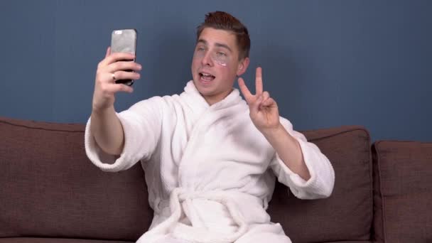 En ung man med fläckar i ansiktet tar en selfie och tar bort fläckar från ansiktet. Blå hydrogelplåster för ansiktsföryngring. En man sitter i en vit rock på soffan. — Stockvideo