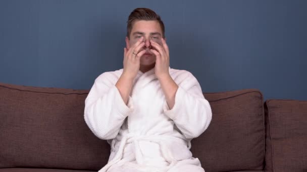 Um jovem com manchas na cara tira uma selfie. Manchas de hidrogel azul para rejuvenescimento facial. Um homem senta-se com um casaco branco no sofá. — Vídeo de Stock