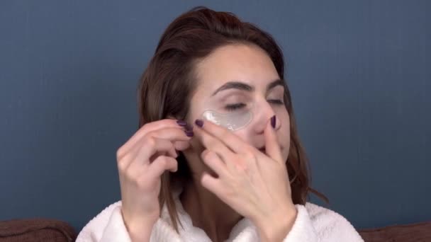 Ung kvinna sätter fläckar på ansiktet. Blå hydrogelplåster för att föryngra huden i ansiktet. Hon sitter i en vit rock. Närbild — Stockvideo