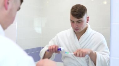 Genç bir adam dişlerini fırçalıyor. Beyaz önlüklü bir adam aynada dişlerine baktı ve onları diş fırçasıyla fırçalamaya başladı. Aynaya bak.