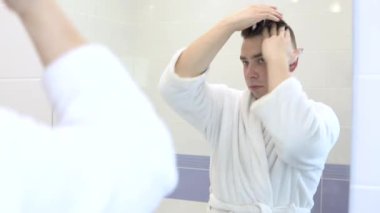 Genç bir adam saçlarını örtmek için saç köpüğü sürüyor. Banyodaki aynanın önünde beyaz bornozlu bir adam. Aynaya bak.