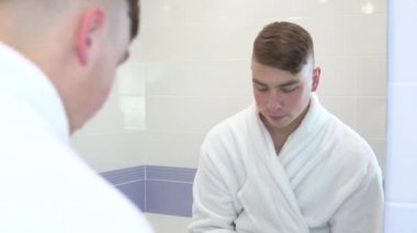 Genç bir adam yüzüne kozmetik bantlar yapıştırıyor. Spa tedavileri. Deri yenilenmesi için kahverengi hidrojel yamalar. Banyodaki aynanın önünde beyaz bir bornozla duruyor. Aynaya bak.