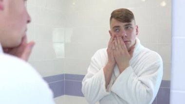 Genç bir adam yüzüne kozmetik bantlar yapıştırıyor. Spa tedavileri. Deri yenilenmesi için kahverengi hidrojel yamalar. Banyodaki aynanın önünde beyaz bir bornozla duruyor. Aynaya bak.