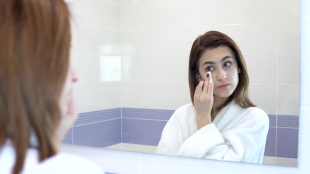 Mladá žena si odstraňuje make-up z obličeje vatou. Dívka v bílém županu před zrcadlem v koupelně. Pohled do zrcadla — Stock video