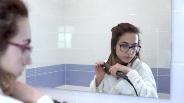 Een jonge vrouw met een bril trekt haar haar recht met een krultang voor een spiegel. Een meisje in een witte jas maakt een kapsel in de badkamer. Zicht door de spiegel — Stockvideo