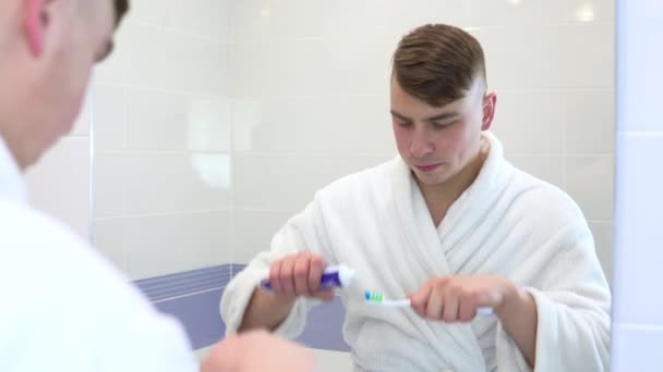 Un jeune homme se brosse les dents. Un homme en manteau blanc regarda ses dents dans le miroir et commença à les brosser avec une brosse à dents. Vue à travers le miroir — Video