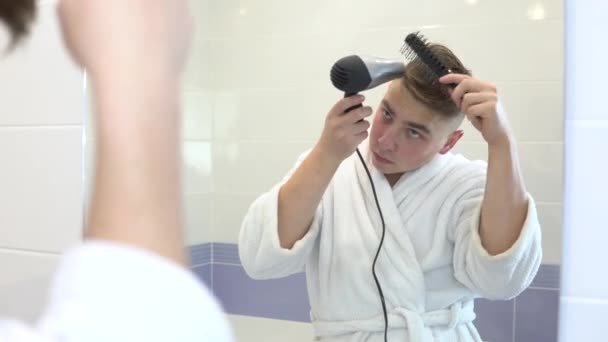 Молодий чоловік сушить волосся феном. Чоловік у білому пальто робить стиль, використовуючи фен і гребінець. Піднімається перед дзеркалом у ванній. Перегляд через дзеркало — стокове відео