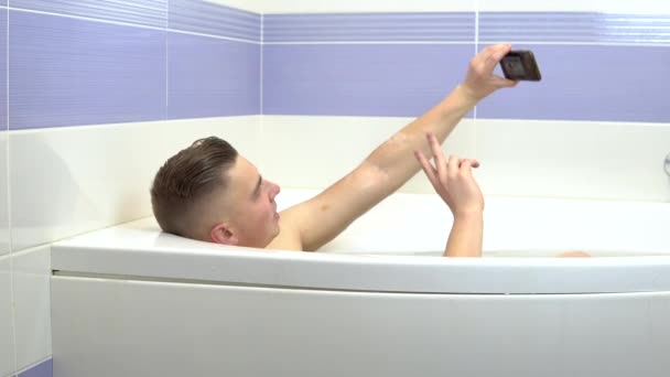 Um jovem deita-se no banho e tira uma selfie ao telefone. Um homem relaxa no banho . — Vídeo de Stock