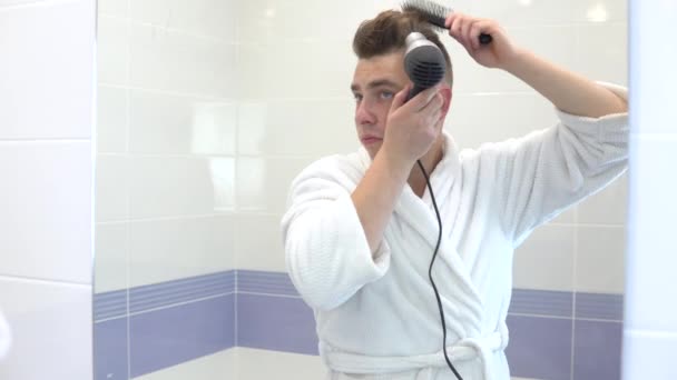 Mladý muž si osušuje vlasy fénem. Muž v bílém plášti dělá styling s fénem a hřebenem. Stojí před zrcadlem v koupelně. Pohled do zrcadla — Stock video