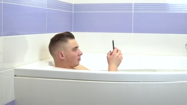 Genç bir adam elinde telefonuyla banyoda yatıyor. Bir adam banyoda rahatlar ve sohbet eder.. — Stok video