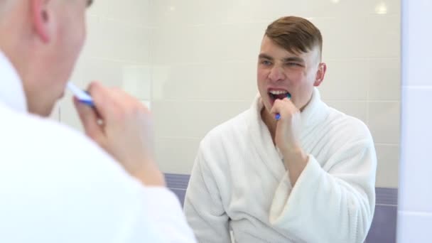 Egy fiatalember aktívan mossa a fogát. Egy férfi fehér fürdőköpenyben a fürdőszobai tükör előtt. Nézz át a tükrön. — Stock videók