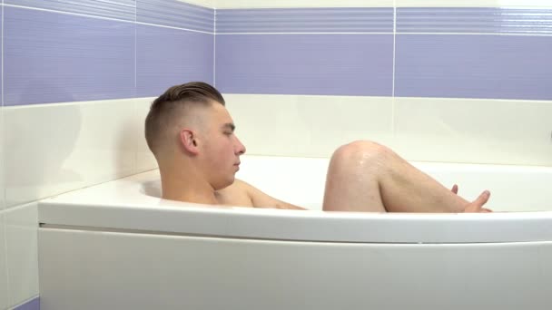 Un joven está acostado en el baño. Un hombre se relaja en el baño y saca su pie del agua . — Vídeos de Stock