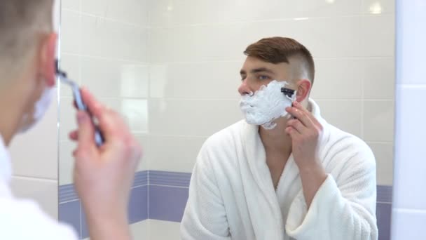 Mladý muž si holí vlasy před zrcadlem. Muž v bílém kabátě s pěnou na obličeji si holí vlasy. Pohled do zrcadla. — Stock video