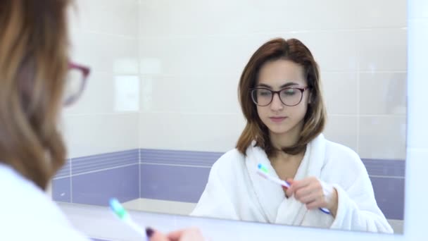 Молодая женщина в очках чистит зубы. Женщина в белом халате начала чистить зубы. Взгляд в зеркало — стоковое видео