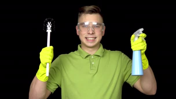 Un joven limpiador sostiene herramientas de limpieza y sonríe. Hombre con gafas de seguridad y guantes para limpiar el inodoro. El tipo sostiene un cepillo de inodoro y spray. Sobre un fondo negro — Vídeo de stock