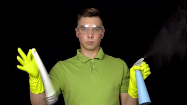 A young man twists a toilet brush on her finger and sprinkles water from a spray bottle. Man in safety glasses and gloves for cleaning the toilet. The guy holds a toilet brush and spray. On a black — Stock Video
