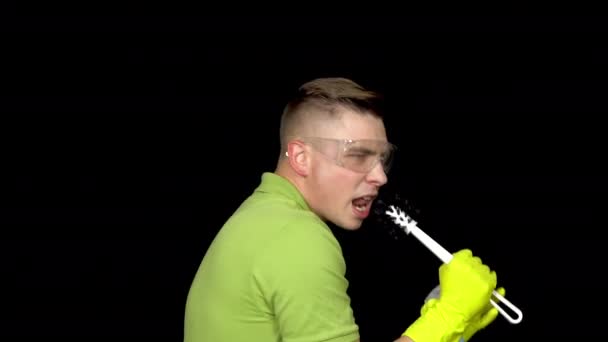 A young man sings in a toilet brush as in a microphone. Man in safety glasses and gloves for cleaning the toilet. The guy holds a toilet brush and spray. On a black background — Stock Video