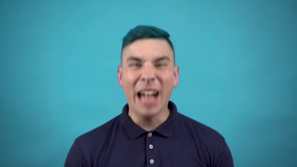 Ein junger Mann mit Brille und blauem Haar schreit vor Wut. Emotionaler Mann schreit auf blauem Hintergrund. — Stockvideo