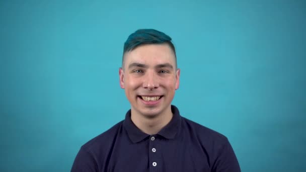 Ein junger Mann mit Brille und blauem Haar lächelt vor Freude. Der emotionale Mann lächelt auf blauem Hintergrund. — Stockvideo