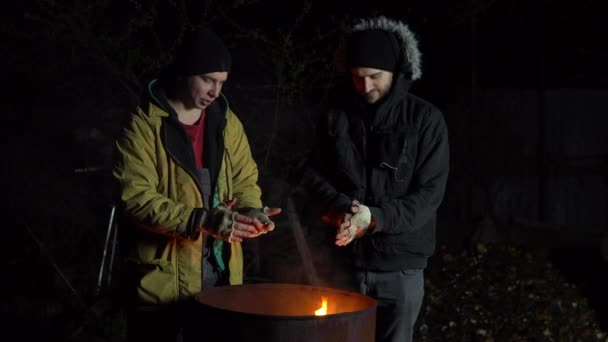 Zwei obdachlose junge Männer sonnen sich im Feuer. Männer stehen nachts in der Nähe einer Feuertonne. — Stockvideo