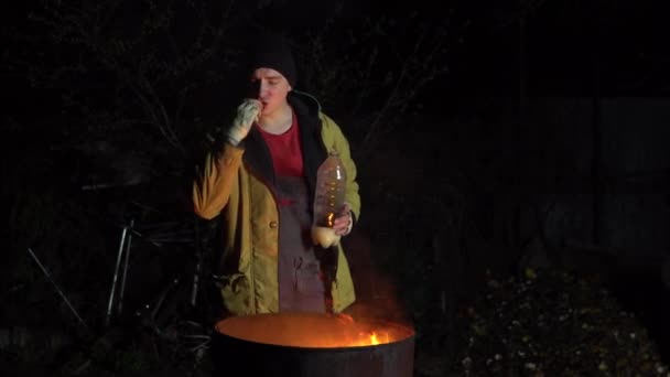En hemlös ung man står vid elden och äter ett äpple. En man står på natten nära en tunna eld och solfjäder. — Stockvideo
