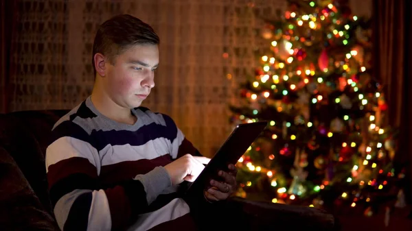 Un joven se sienta con una tableta en las manos sobre el fondo de un árbol de Navidad. La habitación está decorada y llena de ambiente navideño. Tableta electrónica moderna . —  Fotos de Stock