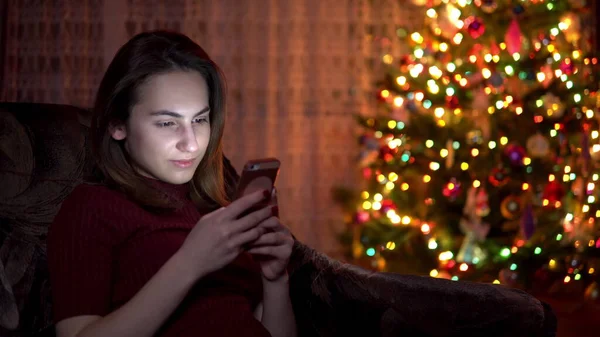 Una joven se sienta con una tableta en las manos sobre el fondo de un árbol de Navidad. La habitación está decorada y llena de ambiente navideño. Tableta electrónica moderna . —  Fotos de Stock
