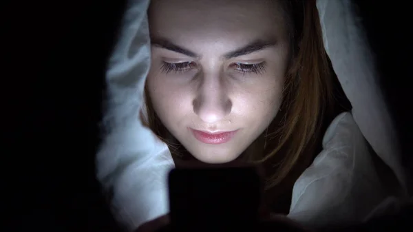 Una mujer joven yace debajo de una manta con un teléfono en sus manos. Una mujer en la oscuridad usa una almohadilla. Tableta electrónica moderna. Primer plano —  Fotos de Stock